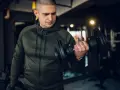Mężczyzna w zielonej bluzie damskiej na siłownię ćwiczy biceps z hantlem.