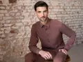 Mężczyzna w brązowej koszuli na wierzchu i spodniach siedzi na tle ceglanej ściany.