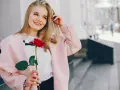 Uśmiechnięta blondynka z różą w dłoni, w różowym płaszczu. Idealna stylizacja: jak się ubrać do kina z chłopakiem, by wyglądać olśniewająco.