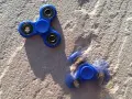 Dwa niebieskie fidget spinnery na piasku, jeden nieruchomy, drugi w ruchu, przypominają czasy, kiedy fidget spinnery były popularne.