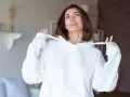 Kobieta w białej bluzie z kapturem, trzymająca za sznurek do bluzy z kapturem, patrzy w górę z uśmiechem.
