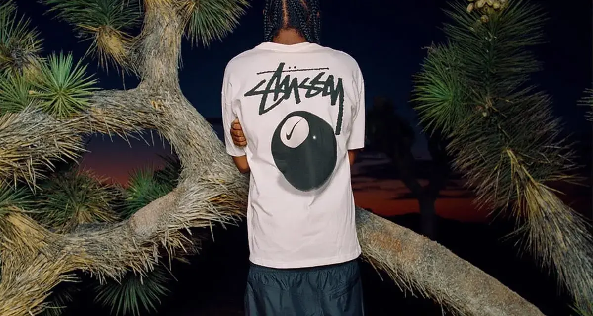 Mężczyzna w koszulce Stussy z grafiką kuli bilardowej 8 i logo Nike. Stussy to kultowa marka streetwearowa.