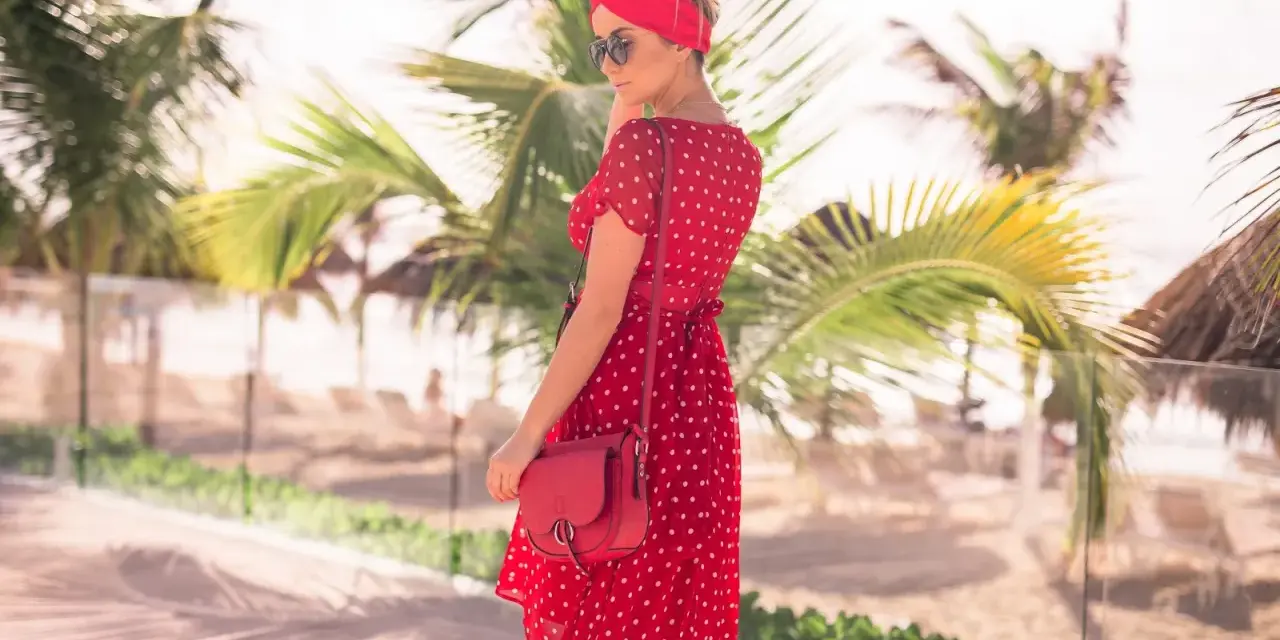 Kobieta w zwiewnej, **czerwonej sukience boho** w białe grochy, z czerwoną opaską na głowie, pozuje na tle palmy i plaży.