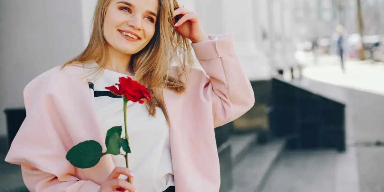 Uśmiechnięta blondynka z różą w dłoni, w różowym płaszczu. Idealna stylizacja: jak się ubrać do kina z chłopakiem, by wyglądać olśniewająco.