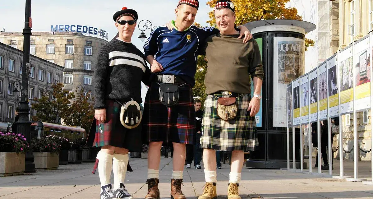 Trzech mężczyzn w tradycyjnych szkockich kiltach pozuje do zdjęcia na tle miejskiej zabudowy. Kto nosi kilt? Ci panowie!