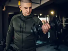 Mężczyzna w zielonej bluzie damskiej na siłownię ćwiczy biceps z hantlem.