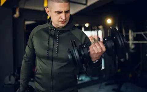 Mężczyzna w zielonej bluzie damskiej na siłownię ćwiczy biceps z hantlem.