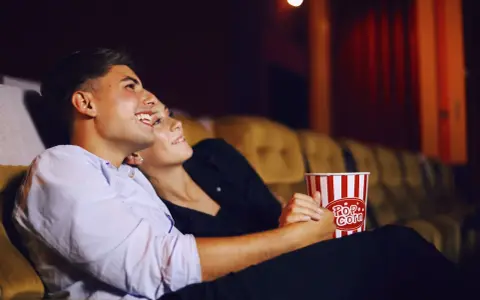 Para randkę do kina wybierz wygodny, ale stylowy strój. Para siedzi blisko siebie, trzymając popcorn.