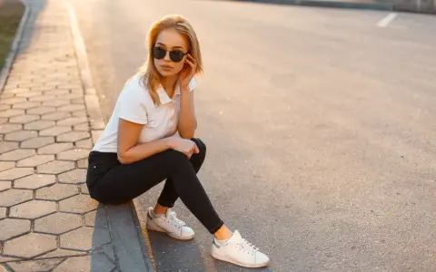Blondynka w czarnych jeansach i białej koszulce polo, siedząca na chodniku. Stylowa inspiracja, jak nosić koszulka polo damska na co dzień.