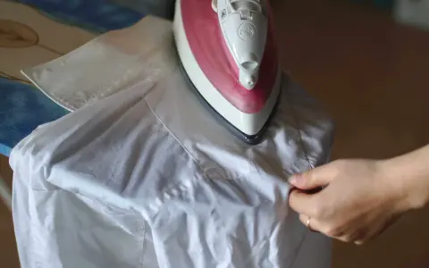 Dłoń układa białą koszulę na desce do prasowania. Czy rękawy koszuli prasuje się na kant? To pytanie zadaje sobie wiele osób podczas prasowania.