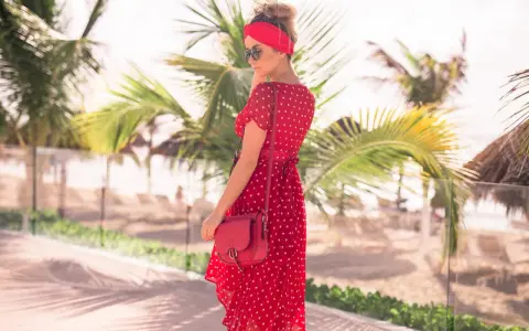 Kobieta w zwiewnej, **czerwonej sukience boho** w białe grochy, z czerwoną opaską na głowie, pozuje na tle palmy i plaży.