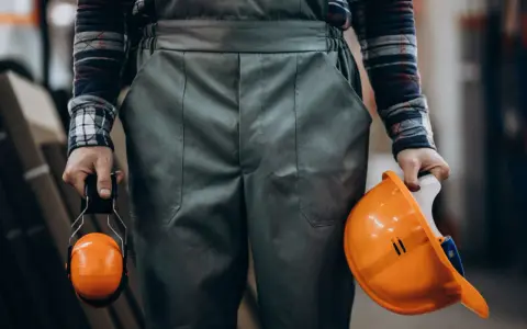 Pracownik w odzieży roboczej trzyma kask i nauszniki. Przepisy dotyczące prania odzieży roboczej są ważne dla bezpieczeństwa.