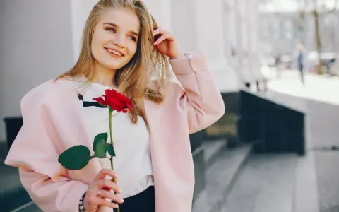 Uśmiechnięta blondynka z różą w dłoni, w różowym płaszczu. Idealna stylizacja: jak się ubrać do kina z chłopakiem, by wyglądać olśniewająco.