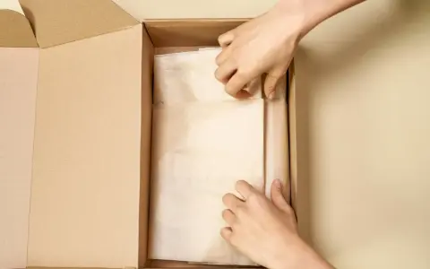 Dłonie układają bluzkę w papierze ozdobnym, przygotowując ją do wysyłki w kartonie.