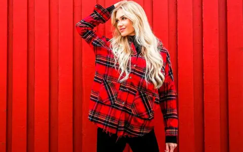 Blondynka w czarnej golfie i modnej koszuli w czerwone paski, pozuje na tle czerwonej, drewnianej ściany.