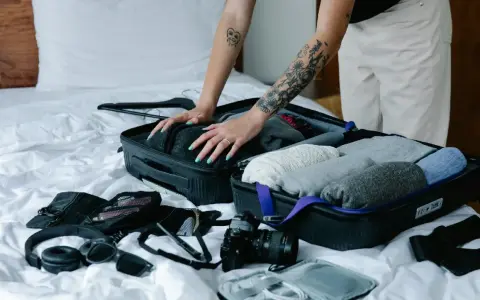 Osoba z tatuażami układa ubrania, pokazując, jak składać bluzy do walizki. Obok leżą słuchawki i aparat.