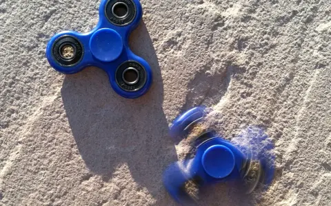 Dwa niebieskie fidget spinnery na piasku, jeden nieruchomy, drugi w ruchu, przypominają czasy, kiedy fidget spinnery były popularne.