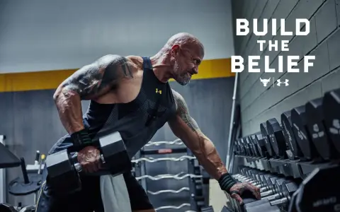 Dwayne "The Rock" Johnson w koszulce Under Armour wykonuje ćwiczenia z hantlami. W tle stojaki z ciężarami.