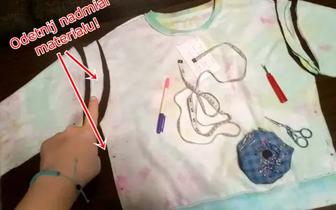 Ręka pokazuje, jak zmniejszyć bluzę, odcinając nadmiar materiału. Na stole leżą miarka, nożyczki i pisak.