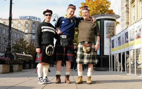 Trzech mężczyzn w tradycyjnych szkockich kiltach pozuje do zdjęcia na tle miejskiej zabudowy. Kto nosi kilt? Ci panowie!