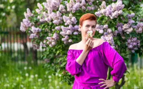 Kobieta w sukience w kolorze fuksja stoi przed kwitnącym bzem, wąchając białe kwiaty.