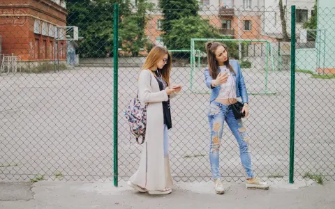 Dwie dziewczyny w modnych stylizacjach, idealnych na outfit do szkoły, stoją przy płocie. Jedna robi sobie selfie, druga przegląda telefon.