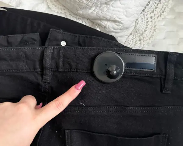 Palec wskazujący z różowym lakierem zbliża się do czarnego klipsa magnetycznego na spodniach. Dowiedz się, jak ściągnąć klips magnetyczny z ubrania.