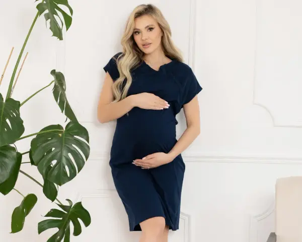 Ciężarna kobieta w granatowej koszuli do porodu, z dłońmi na brzuchu, obok dużej rośliny.