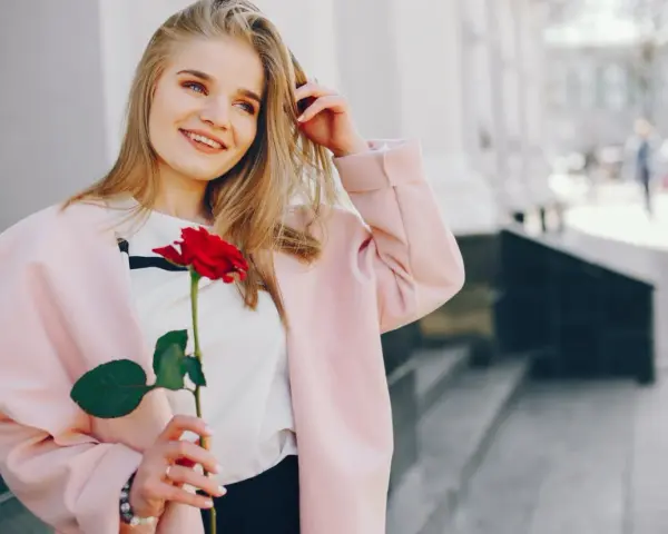 Uśmiechnięta blondynka z różą w dłoni, w różowym płaszczu. Idealna stylizacja: jak się ubrać do kina z chłopakiem, by wyglądać olśniewająco.