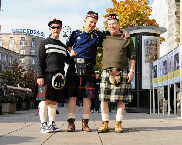 Trzech mężczyzn w tradycyjnych szkockich kiltach pozuje do zdjęcia na tle miejskiej zabudowy. Kto nosi kilt? Ci panowie!