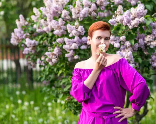 Kobieta w sukience w kolorze fuksja stoi przed kwitnącym bzem, wąchając białe kwiaty.