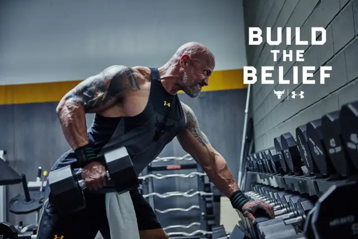 Dwayne "The Rock" Johnson w koszulce Under Armour wykonuje ćwiczenia z hantlami. W tle stojaki z ciężarami.