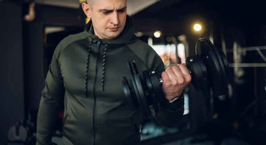 Mężczyzna w zielonej bluzie damskiej na siłownię ćwiczy biceps z hantlem.