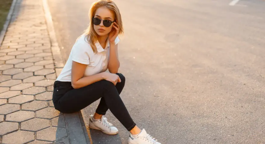 Blondynka w czarnych jeansach i białej koszulce polo, siedząca na chodniku. Stylowa inspiracja, jak nosić koszulka polo damska na co dzień.