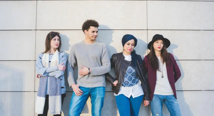 Cztery osoby w różnorodnych stylizacjach stoją przy ścianie. Pokazują, że wszystko może być modne, od casualowych jeansów po eleganckie nakrycia głowy.