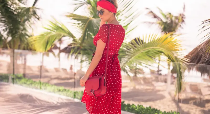 Kobieta w zwiewnej, **czerwonej sukience boho** w białe grochy, z czerwoną opaską na głowie, pozuje na tle palmy i plaży.