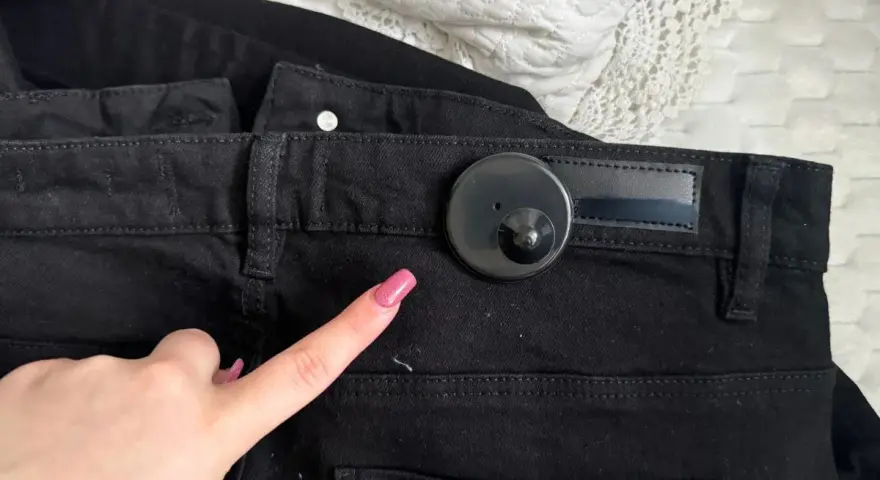 Palec wskazujący z różowym lakierem zbliża się do czarnego klipsa magnetycznego na spodniach. Dowiedz się, jak ściągnąć klips magnetyczny z ubrania.