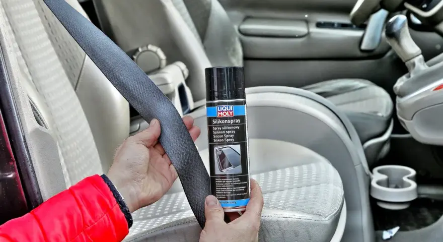 Ręce trzymają pas bezpieczeństwa i puszkę z napisem "Silikonspray". Dowiedz się, jak usunąć silikon z materiału w samochodzie.