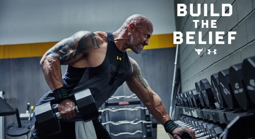Dwayne "The Rock" Johnson w koszulce Under Armour wykonuje ćwiczenia z hantlami. W tle stojaki z ciężarami.