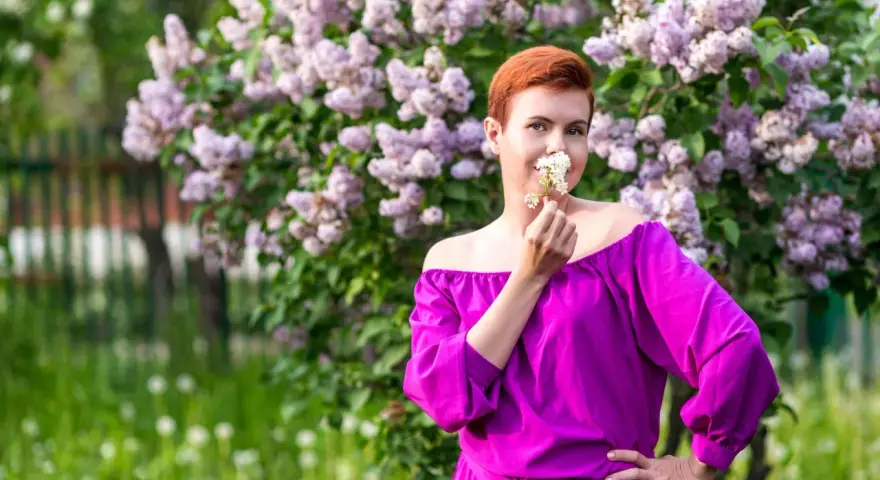 Kobieta w sukience w kolorze fuksja stoi przed kwitnącym bzem, wąchając białe kwiaty.
