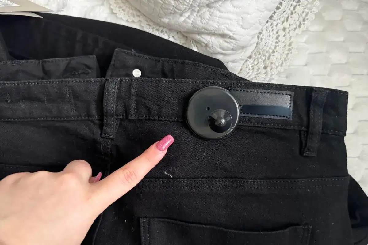 Dłoń z różowym lakierem wskazuje na klips magnetyczny na czarnych spodniach. Jak ściągnąć klips magnetyczny z ubrania?