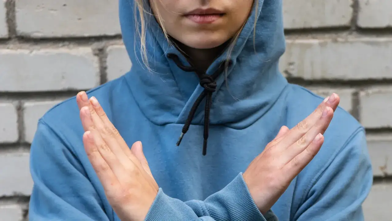 Osoba w niebieskiej bluzie z kapturem krzyżuje ręce. Sznurki od bluzy są zawiązane w supeł.